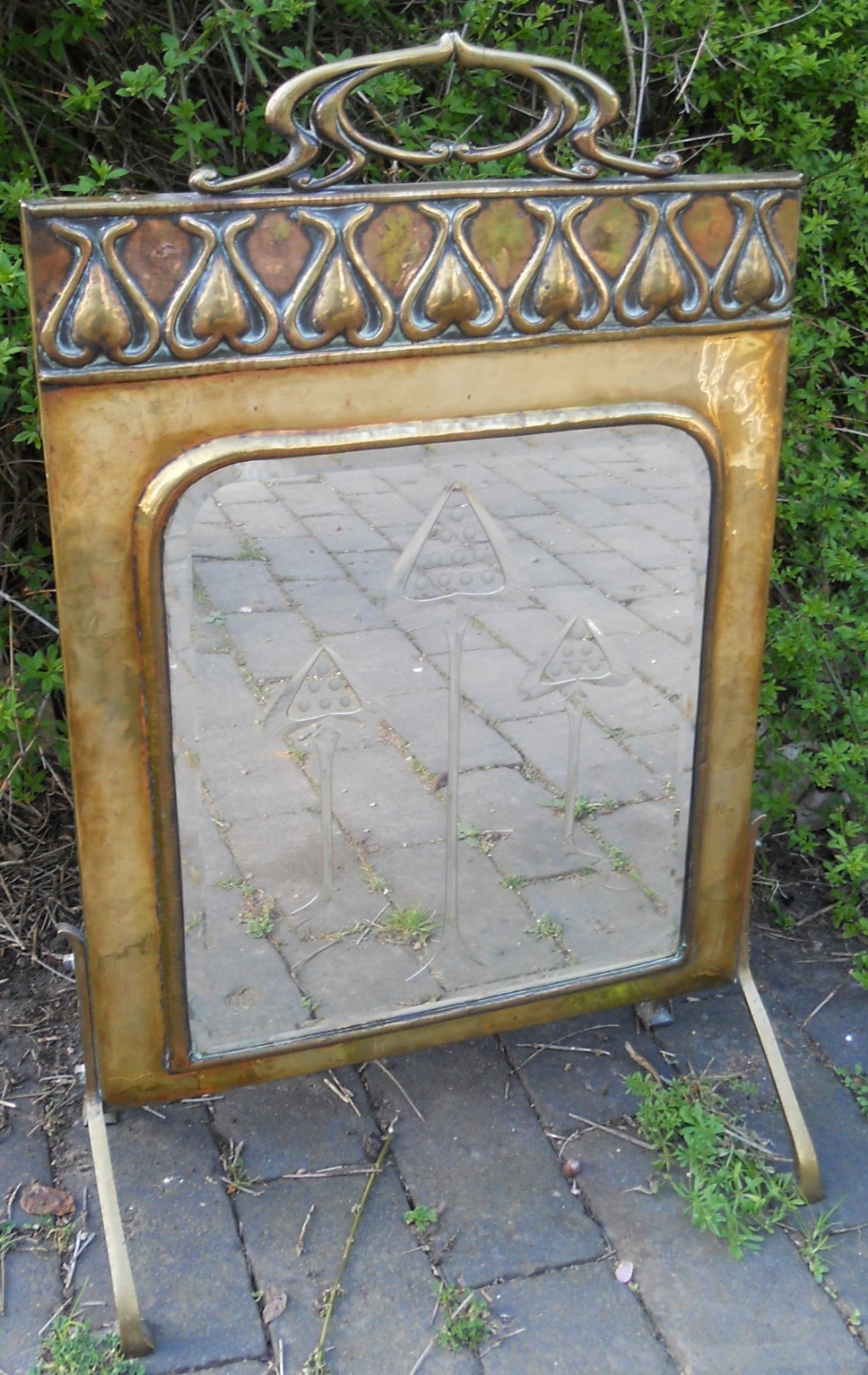 An Art Nouveau brass firescreen featuring an embossed top frieze, curved carrying handle, and a mirrored glass insert etched with stylised tree motifs.