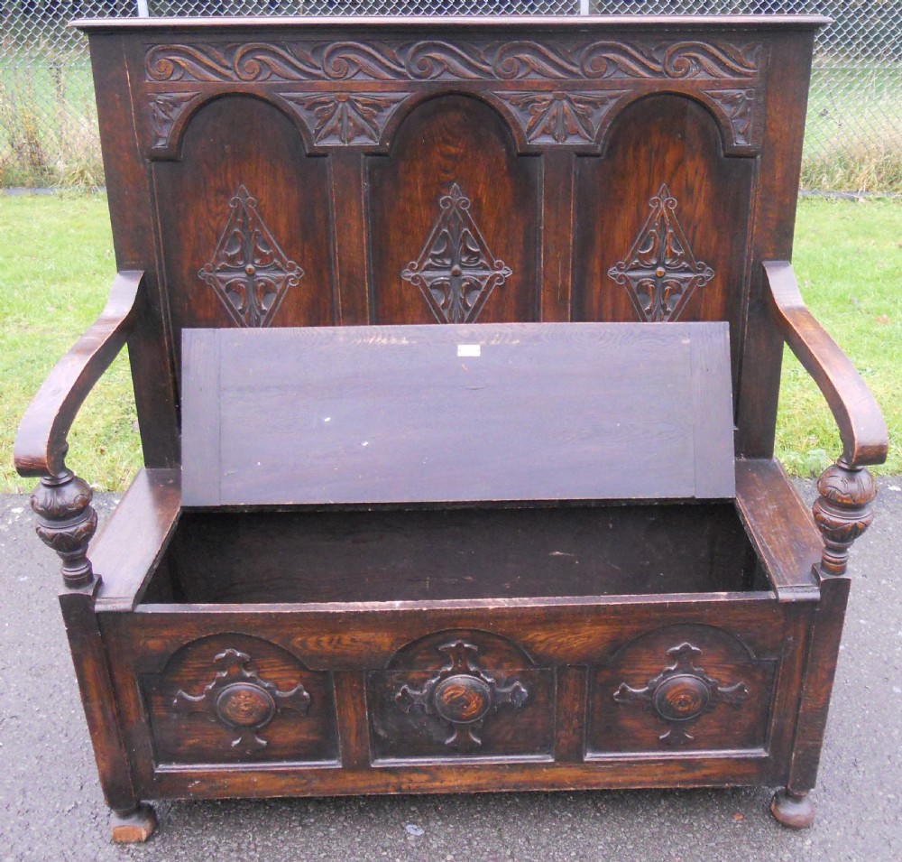 An Edwardian carved dark oak hall settle featuring a high panelled back with decorative arches, carved diamond motifs, turned arm supports, and a hinged seat base. View VI