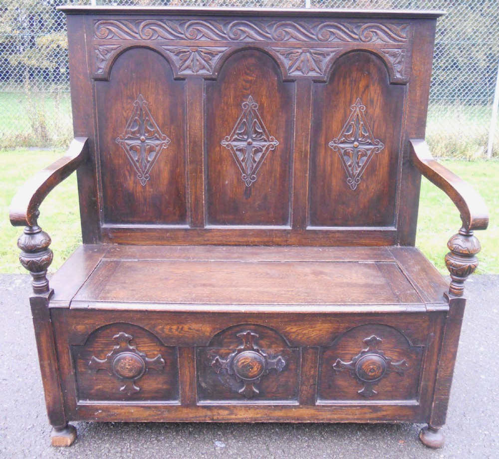 An Edwardian carved dark oak hall settle featuring a high panelled back with decorative arches, carved diamond motifs, turned arm supports, and a hinged seat base. View VII