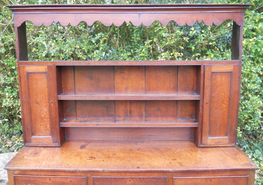A Georgian oak kitchen dresser featuring three base drawers, cabriole front legs, brass handles, and an upper rack with shelves flanked by cupboard doors. View VI