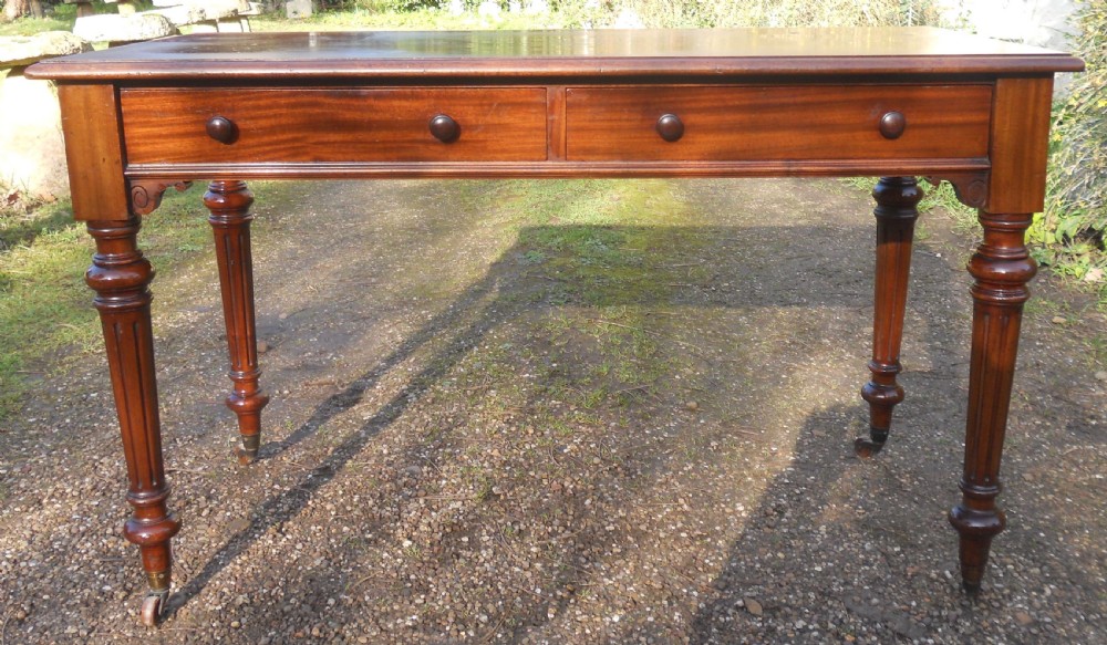 Victorian mahogany writing table featuring two frieze drawers with wooden knob handles, raised on turned and fluted legs with brass castors.