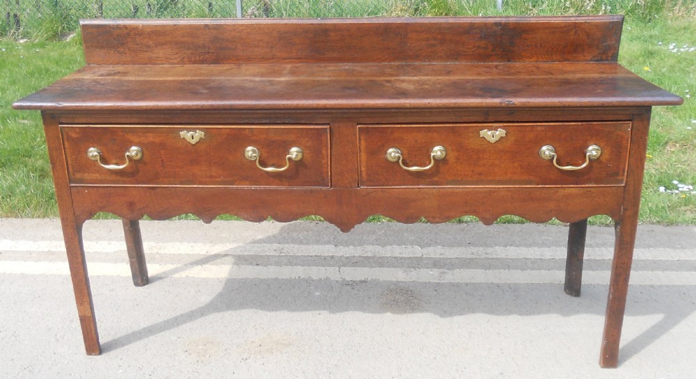A Georgian oak dresser base with two wide drawers, brass swan-neck handles, keyhole escutcheons, a scalloped apron, and a wooden backboard.