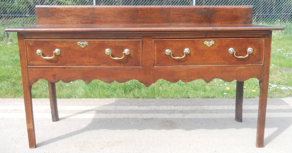 A Georgian oak dresser base with two wide drawers, brass swan-neck handles, keyhole escutcheons, a scalloped apron, and a wooden backboard. View II