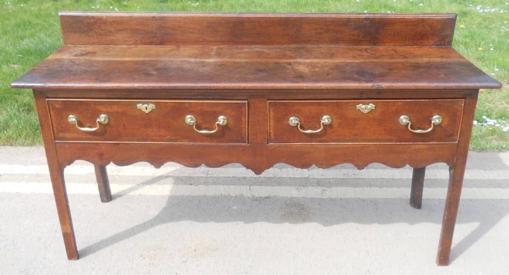 A Georgian oak dresser base with two wide drawers, brass swan-neck handles, keyhole escutcheons, a scalloped apron, and a wooden backboard. View III