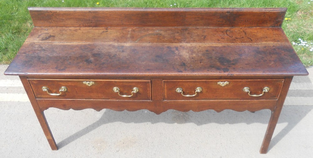 A Georgian oak dresser base with two wide drawers, brass swan-neck handles, keyhole escutcheons, a scalloped apron, and a wooden backboard. View IV