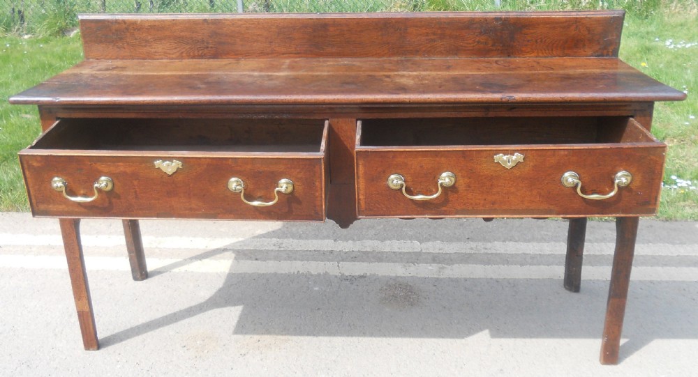 A Georgian oak dresser base with two wide drawers, brass swan-neck handles, keyhole escutcheons, a scalloped apron, and a wooden backboard. View VII