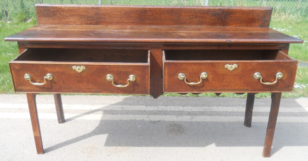 A Georgian oak dresser base with two wide drawers, brass swan-neck handles, keyhole escutcheons, a scalloped apron, and a wooden backboard. View X