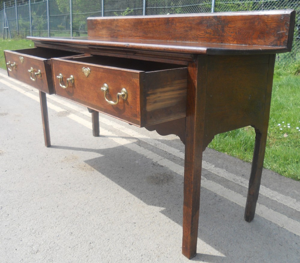 A Georgian oak dresser base with two wide drawers, brass swan-neck handles, keyhole escutcheons, a scalloped apron, and a wooden backboard. View XI