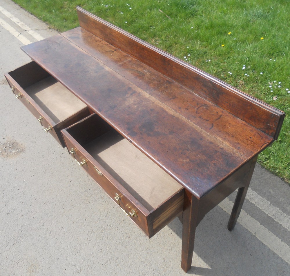 A Georgian oak dresser base with two wide drawers, brass swan-neck handles, keyhole escutcheons, a scalloped apron, and a wooden backboard. View XII
