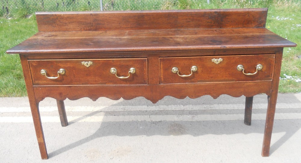 A Georgian oak dresser base with two wide drawers, brass swan-neck handles, keyhole escutcheons, a scalloped apron, and a wooden backboard. View XIII