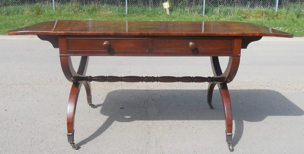 A Regency mahogany sofa table with two drop-leaf ends, twin drawers, turned stretchers, and curved sabre legs on brass castors. View VI