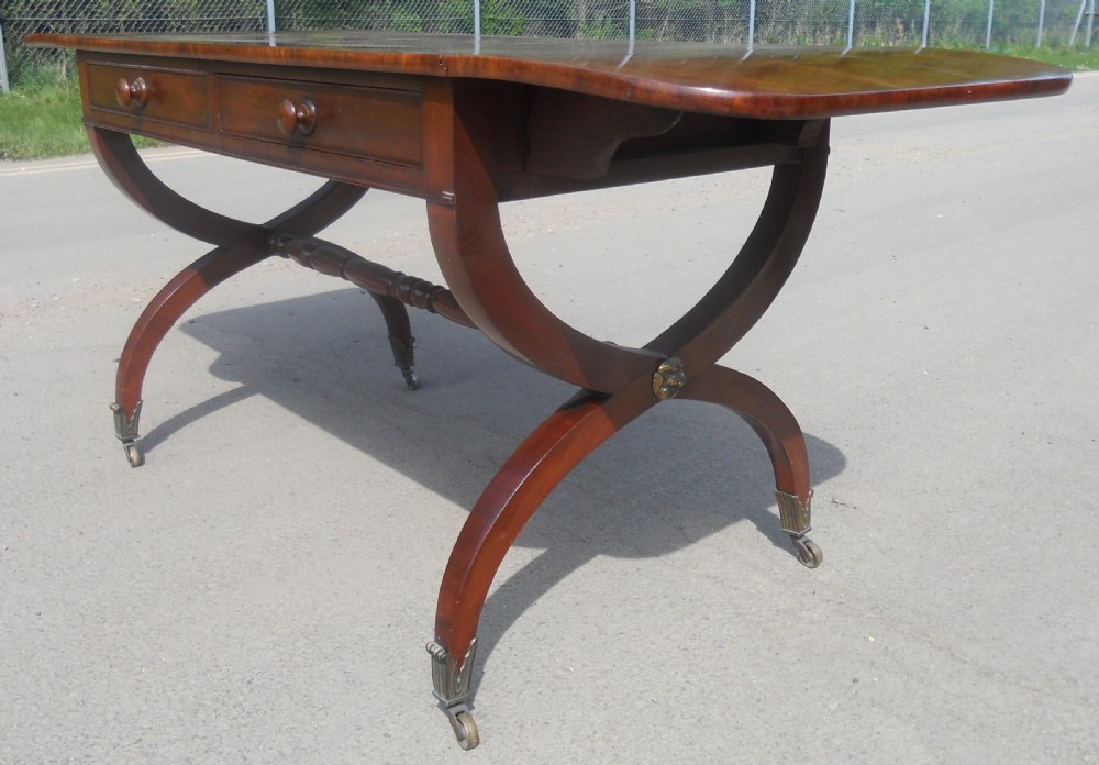 A Regency mahogany sofa table with two drop-leaf ends, twin drawers, turned stretchers, and curved sabre legs on brass castors. View IX