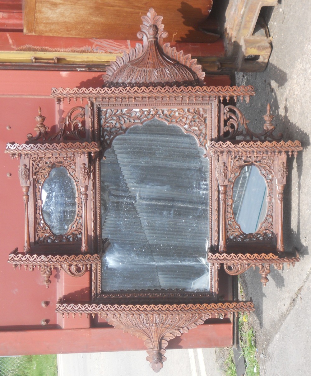 An intricately carved Victorian mahogany wall mirror featuring openwork fretwork, side display shelves with small mirror panels, an arched central mirror, and a carved top pediment.