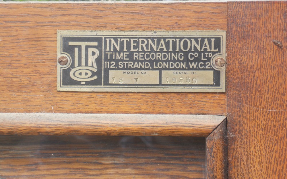 An Edwardian oak-cased factory electric master clock featuring a long glass front panel, a circular white dial with Arabic numerals, and a detached brass pendulum. View VI