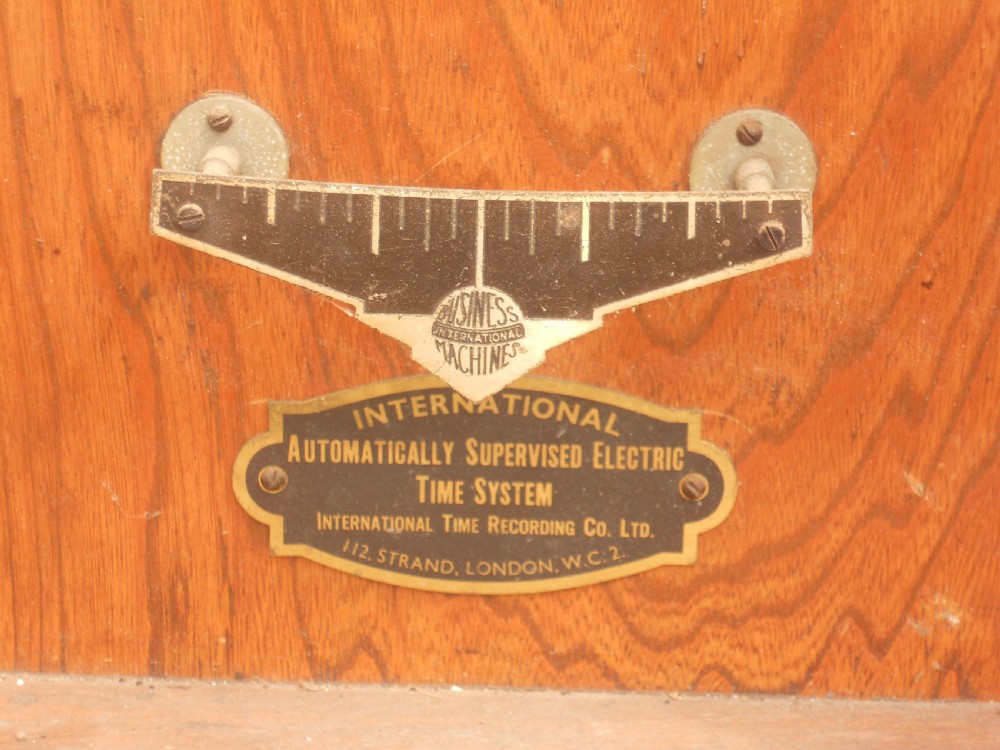An Edwardian oak-cased factory electric master clock featuring a long glass front panel, a circular white dial with Arabic numerals, and a detached brass pendulum. View VII