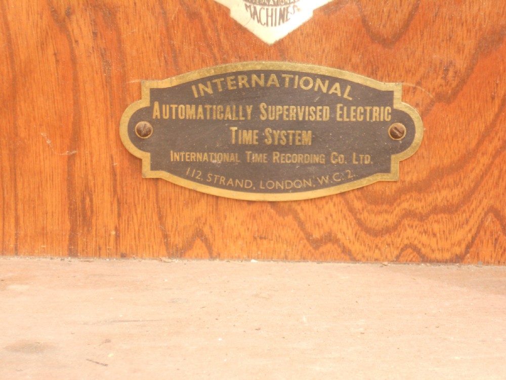 An Edwardian oak-cased factory electric master clock featuring a long glass front panel, a circular white dial with Arabic numerals, and a detached brass pendulum. View VIII
