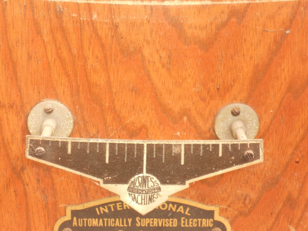 An Edwardian oak-cased factory electric master clock featuring a long glass front panel, a circular white dial with Arabic numerals, and a detached brass pendulum. View IX
