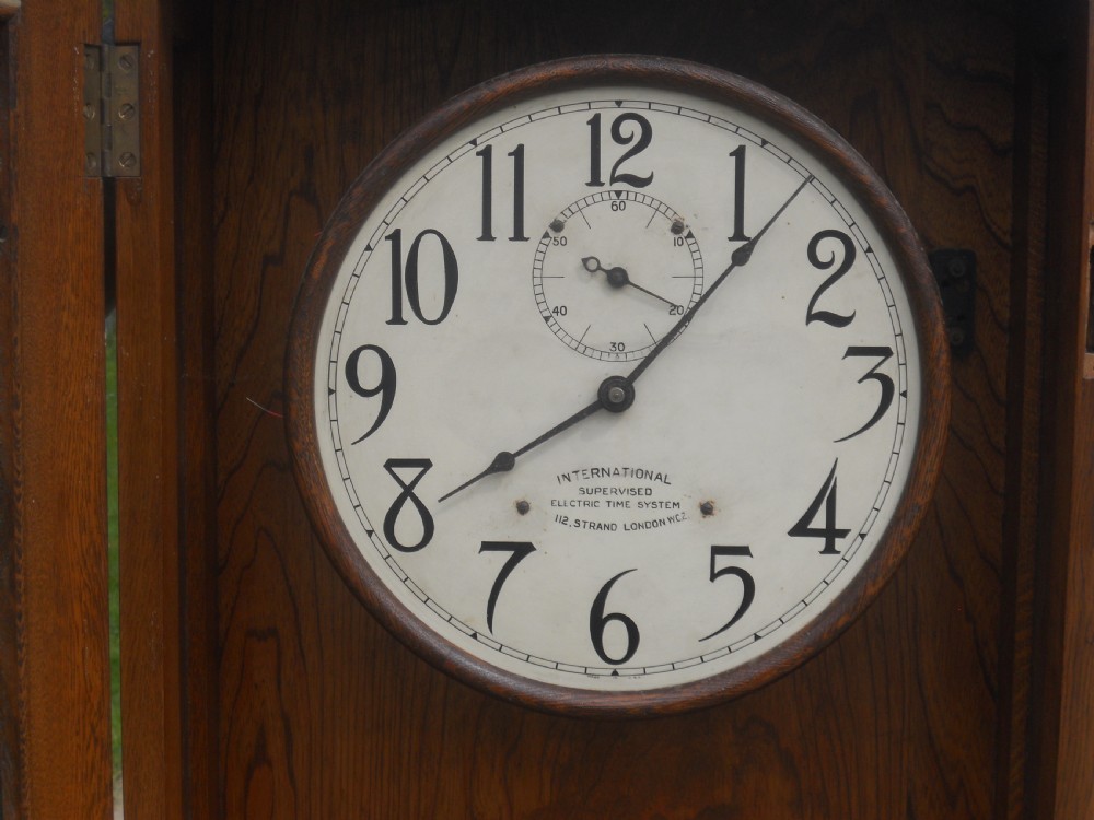 An Edwardian oak-cased factory electric master clock featuring a long glass front panel, a circular white dial with Arabic numerals, and a detached brass pendulum. View X
