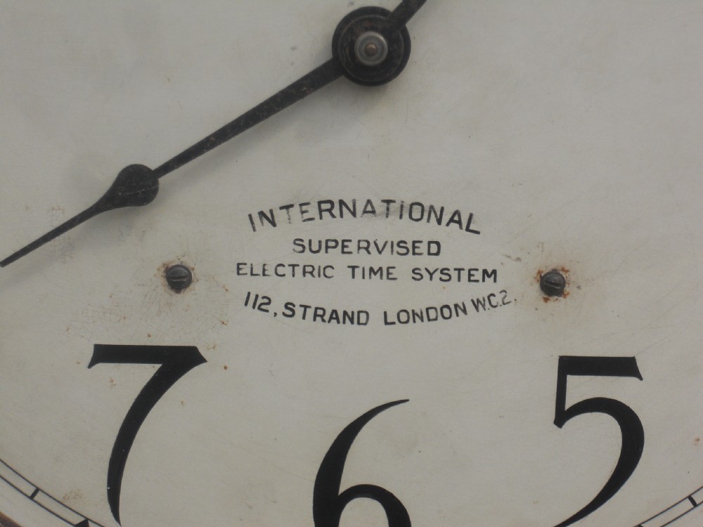 An Edwardian oak-cased factory electric master clock featuring a long glass front panel, a circular white dial with Arabic numerals, and a detached brass pendulum. View XI