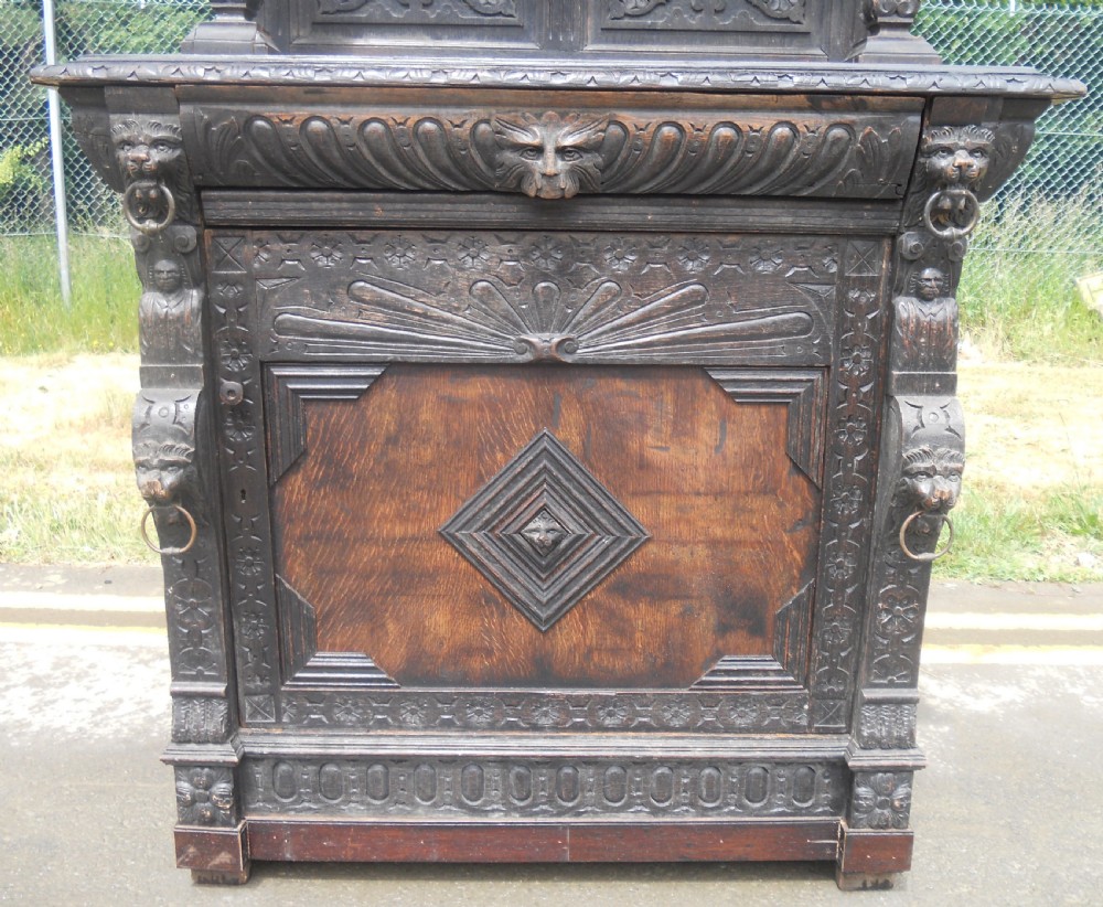 A dark Victorian carved oak side cupboard featuring an upper open shelf supported by sculpted figures, ornate carved masks, and geometric door panel motifs. View IV