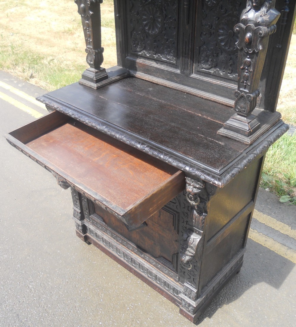 A dark Victorian carved oak side cupboard featuring an upper open shelf supported by sculpted figures, ornate carved masks, and geometric door panel motifs. View VI