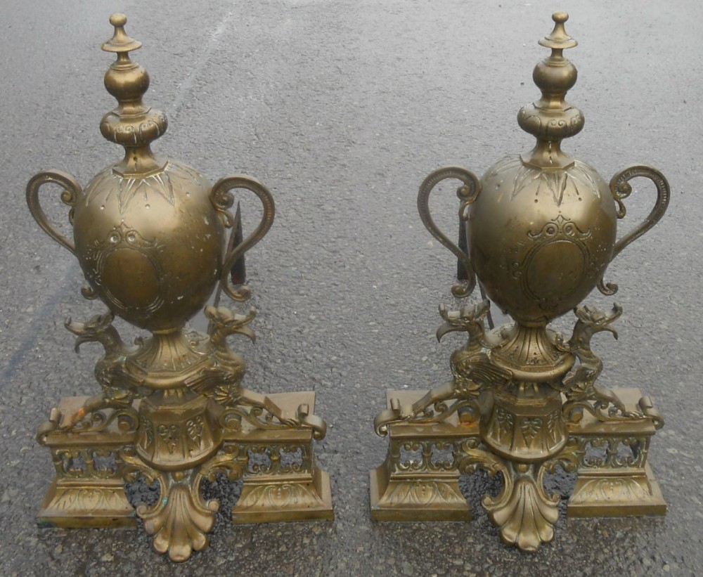 A pair of ornate cast brass fire dogs featuring urn-shaped finials, twin scroll handles, and pierced scrollwork bases.