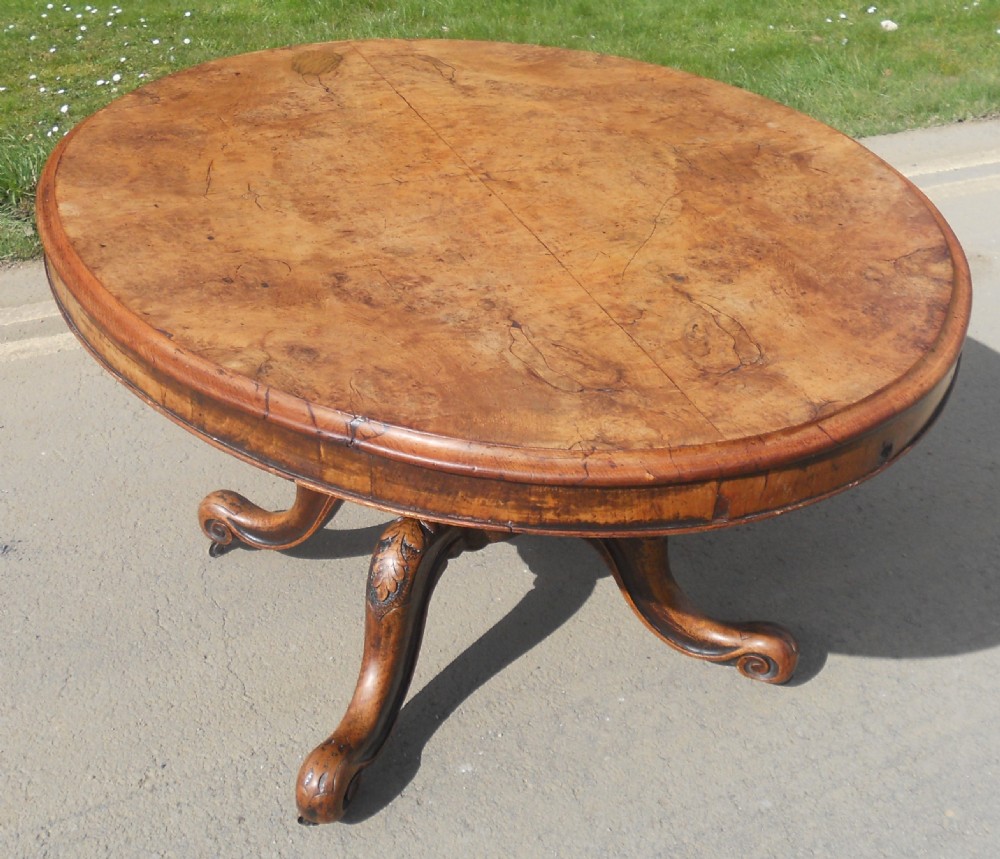 An oval Victorian centre table featuring a figured burr walnut top, carved knees with leaf details, and four scroll-foot legs on castors.