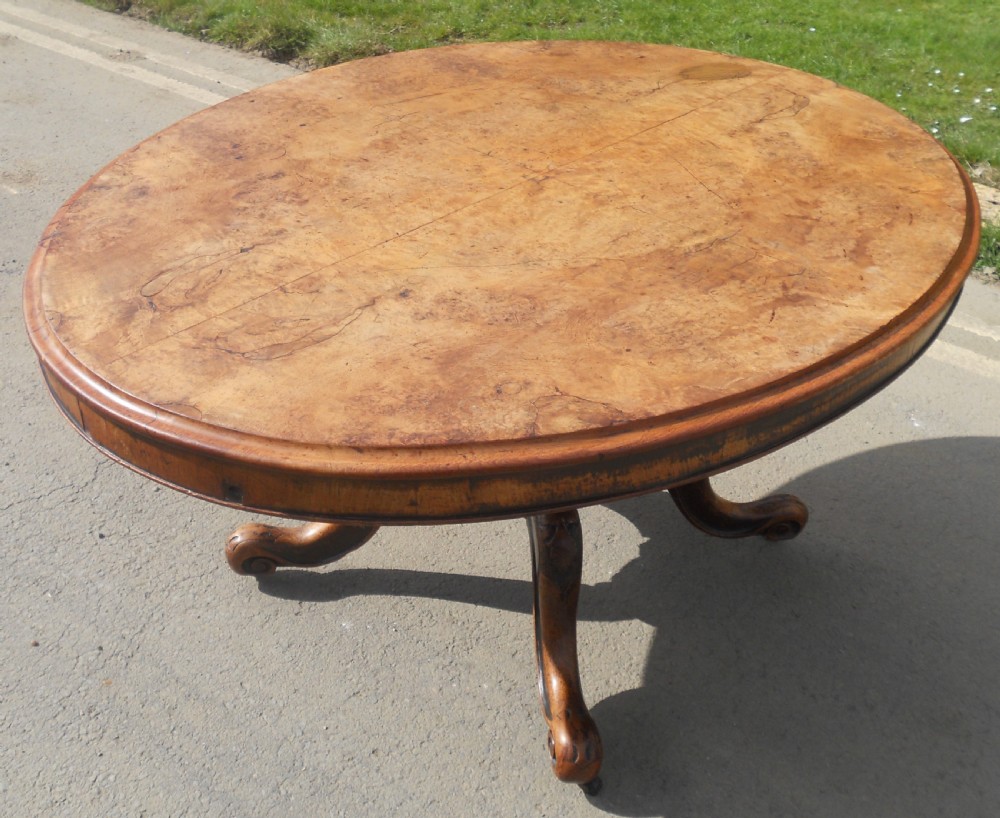 An oval Victorian centre table featuring a figured burr walnut top, carved knees with leaf details, and four scroll-foot legs on castors. View II