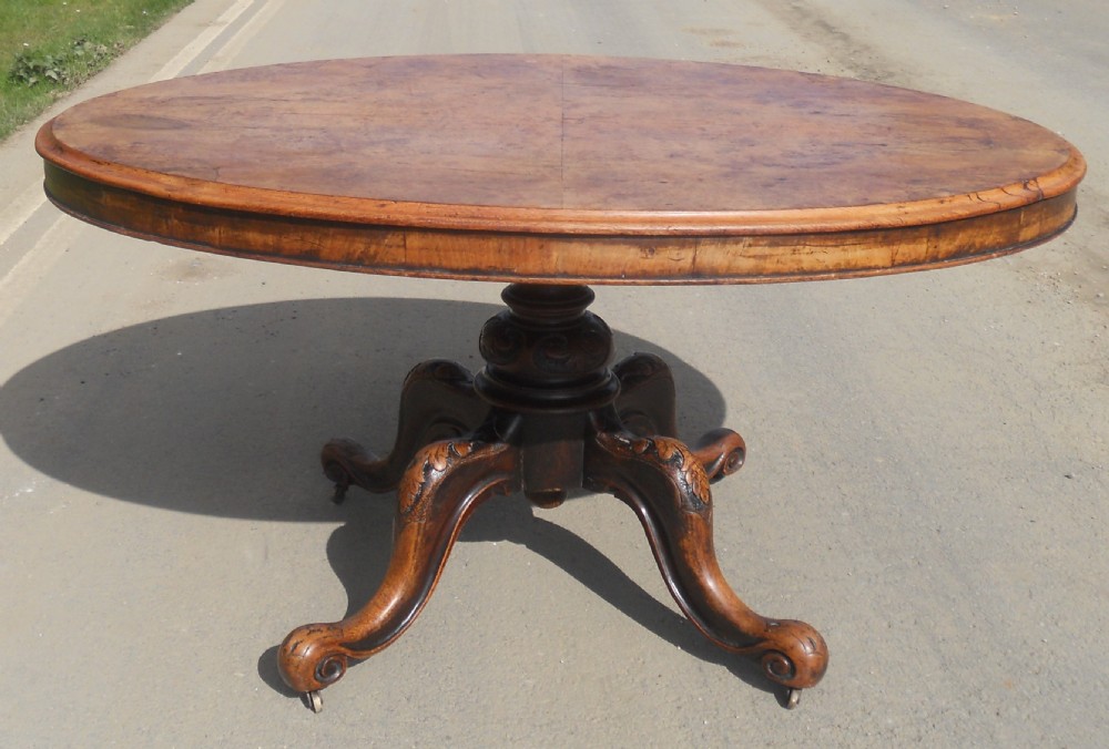 An oval Victorian centre table featuring a figured burr walnut top, carved knees with leaf details, and four scroll-foot legs on castors. View III