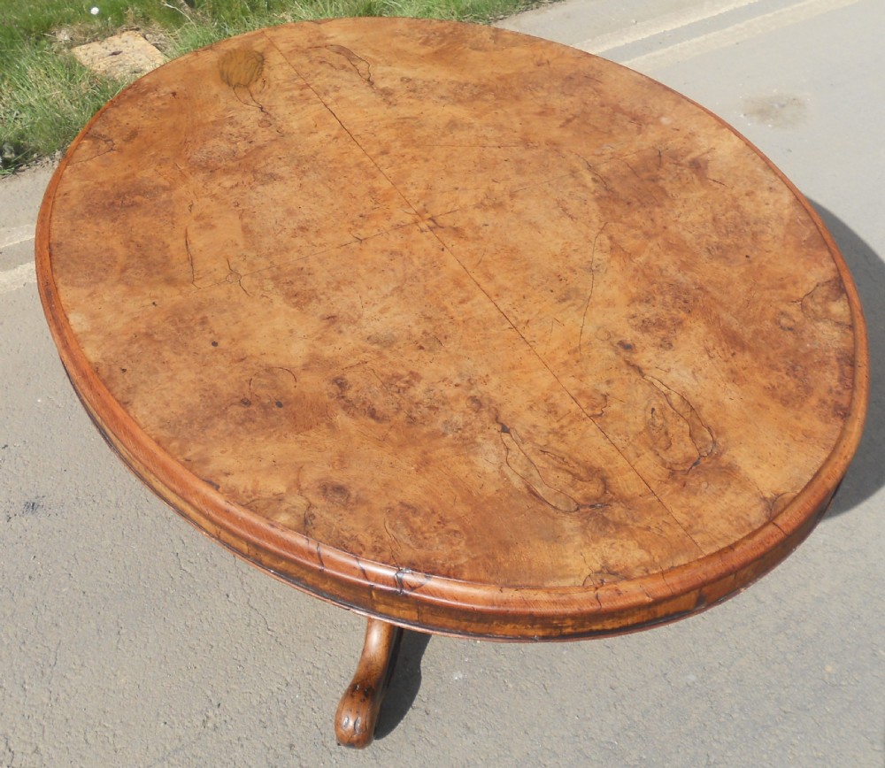 An oval Victorian centre table featuring a figured burr walnut top, carved knees with leaf details, and four scroll-foot legs on castors. View IV