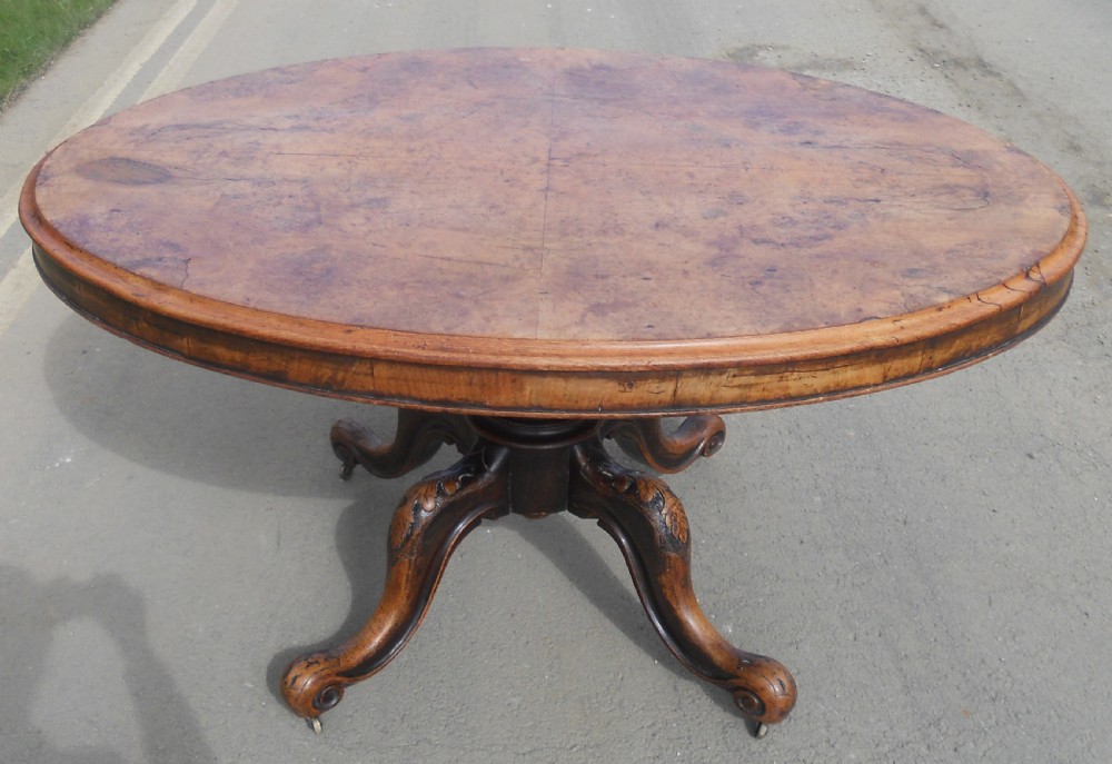 An oval Victorian centre table featuring a figured burr walnut top, carved knees with leaf details, and four scroll-foot legs on castors. View V