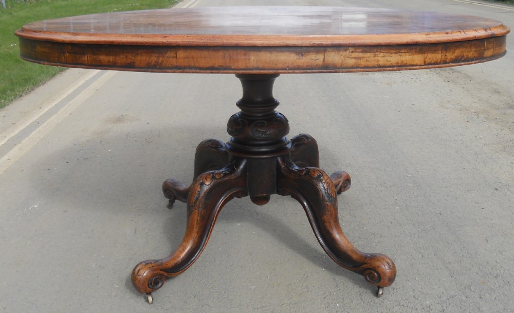 An oval Victorian centre table featuring a figured burr walnut top, carved knees with leaf details, and four scroll-foot legs on castors. View VI
