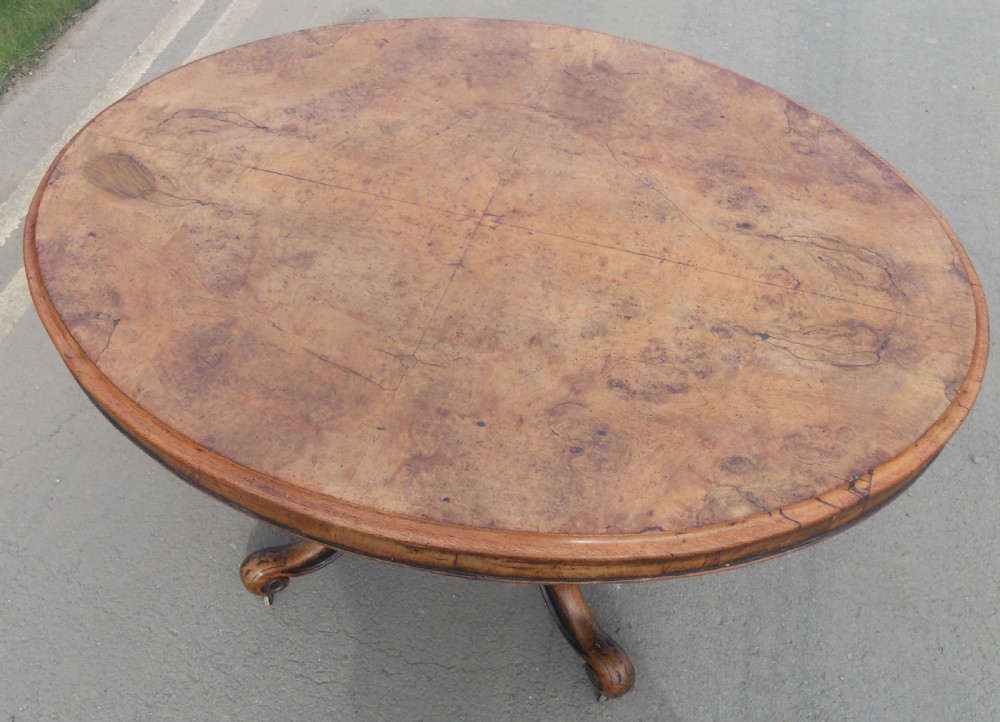An oval Victorian centre table featuring a figured burr walnut top, carved knees with leaf details, and four scroll-foot legs on castors. View VII