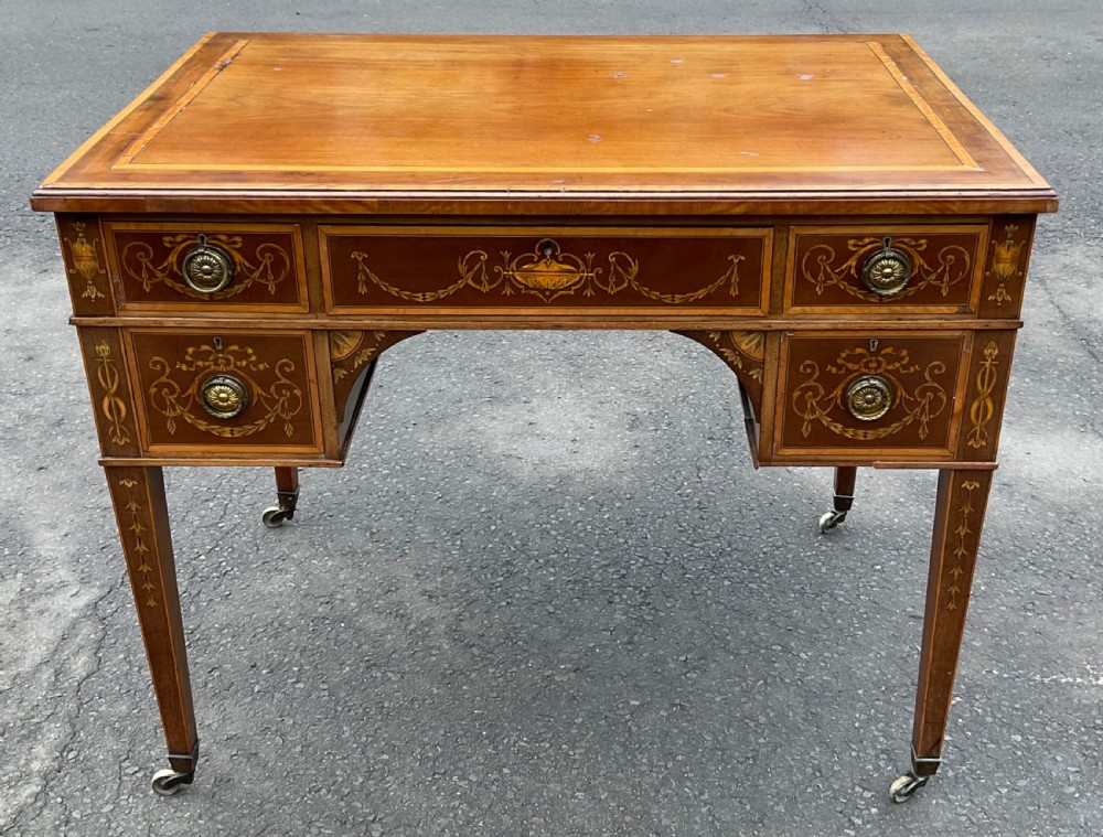 An Edwardian mahogany writing desk featuring urn and floral marquetry inlays, five drawers with circular brass pulls, tapered legs, and brass castors.