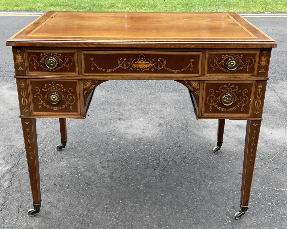An Edwardian mahogany writing desk featuring urn and floral marquetry inlays, five drawers with circular brass pulls, tapered legs, and brass castors. View IV