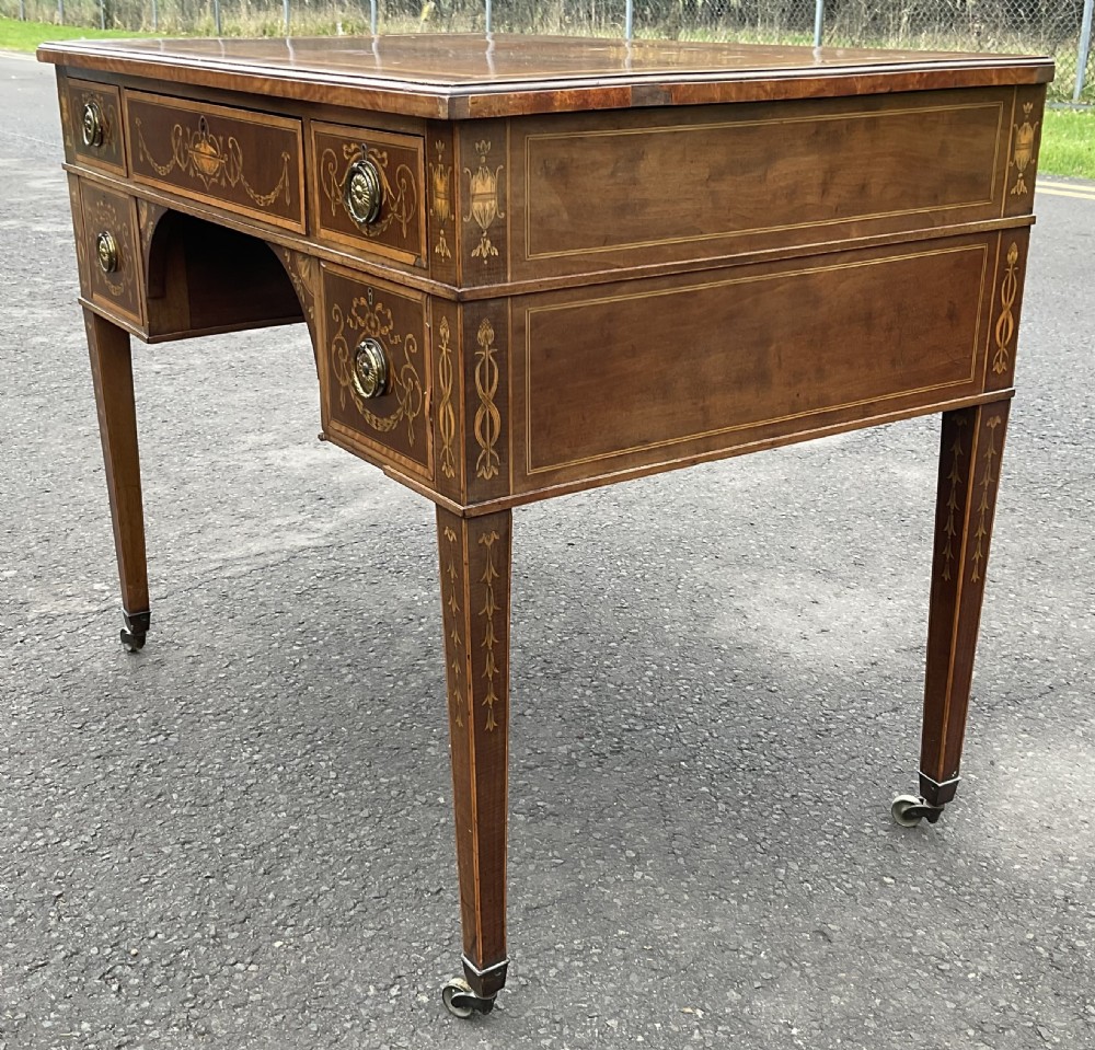 An Edwardian mahogany writing desk featuring urn and floral marquetry inlays, five drawers with circular brass pulls, tapered legs, and brass castors. View V