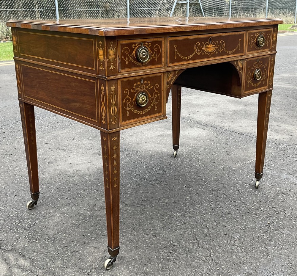 An Edwardian mahogany writing desk featuring urn and floral marquetry inlays, five drawers with circular brass pulls, tapered legs, and brass castors. View VI