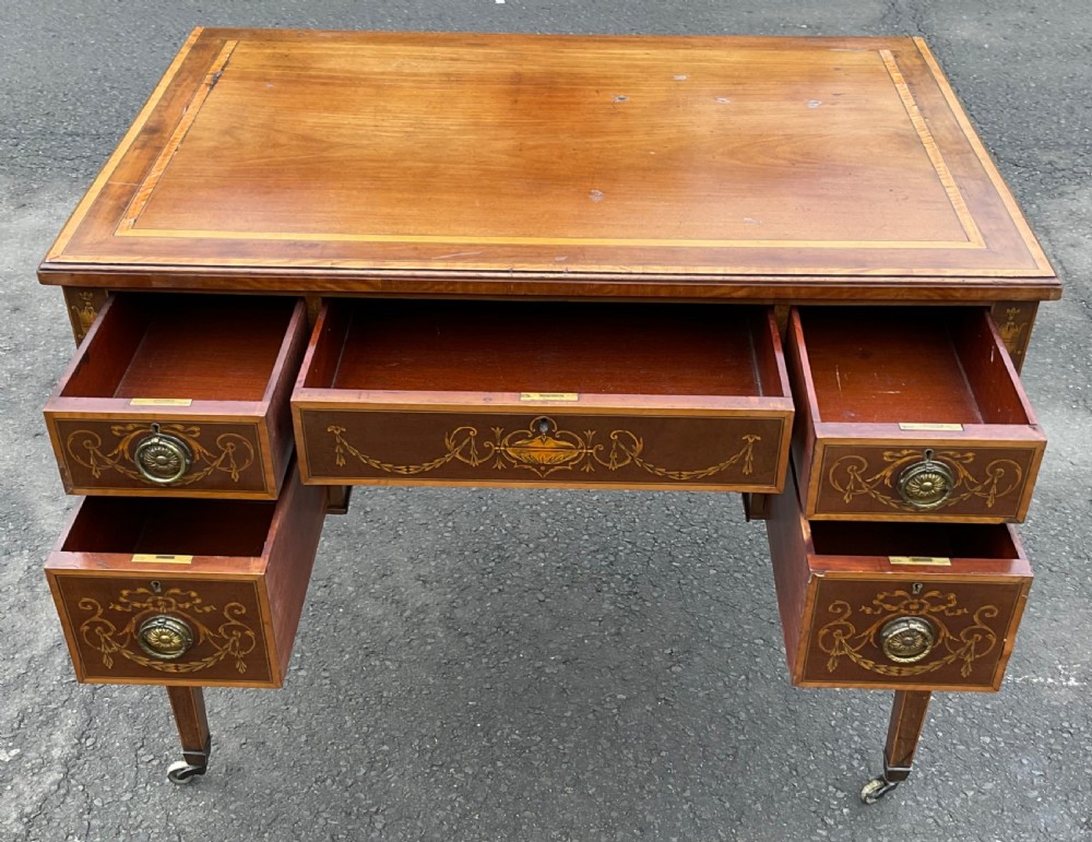 An Edwardian mahogany writing desk featuring urn and floral marquetry inlays, five drawers with circular brass pulls, tapered legs, and brass castors. View X