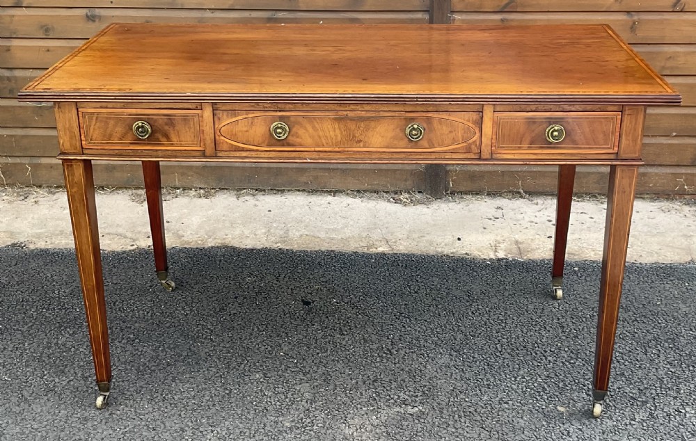An Edwardian mahogany writing table featuring three inlaid drawers, brass ring handles, and square tapered legs on castors.