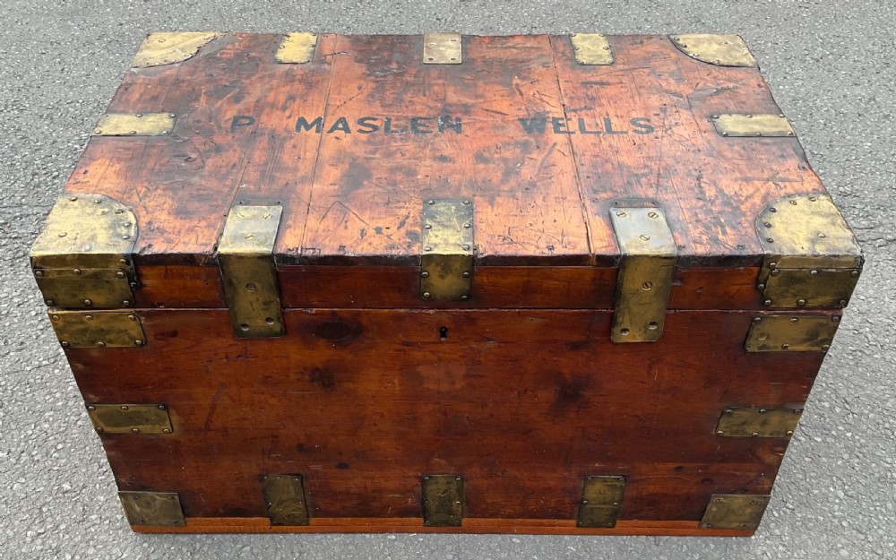 A warm brown wooden military trunk reinforced with brass corner straps, featuring stencilled black lettering reading P. MASLEN WELLS across the top lid.