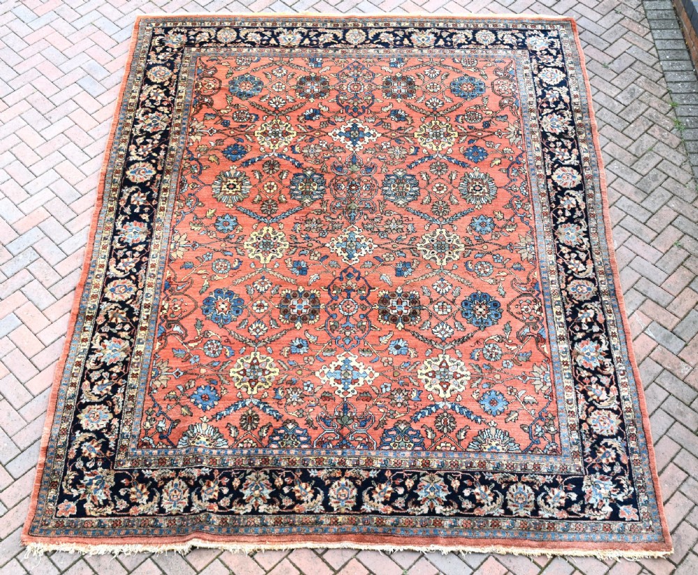 An antique Persian Lilihan wool carpet featuring a terracotta pink field woven with blue floral motifs and framed by a dark navy ornate border.
