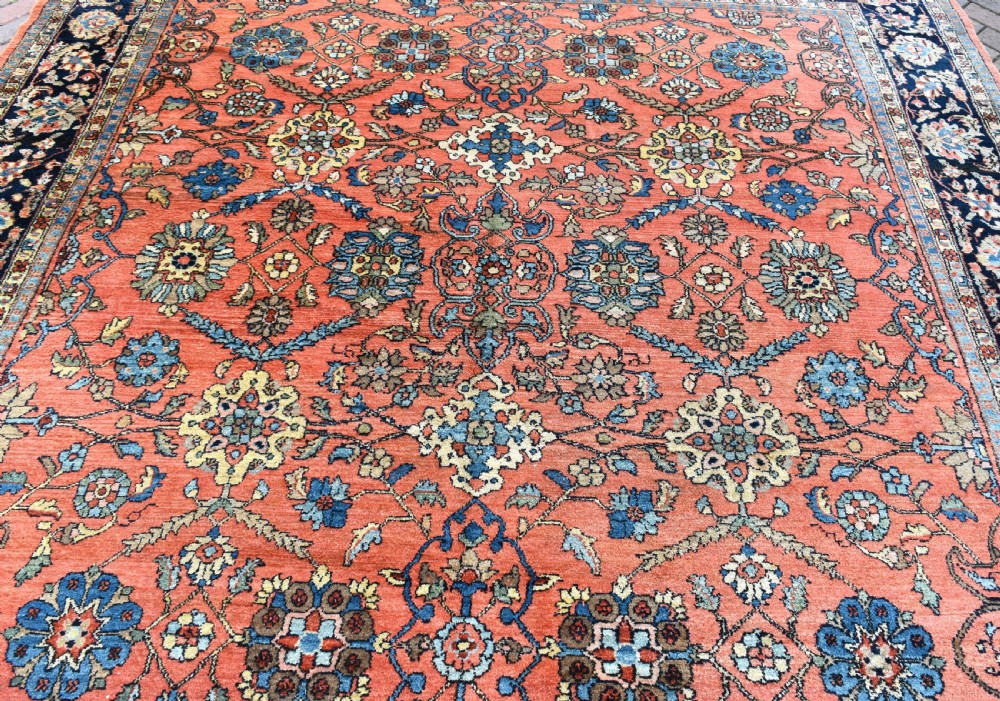 An antique Persian Lilihan wool carpet featuring a terracotta pink field woven with blue floral motifs and framed by a dark navy ornate border. View II