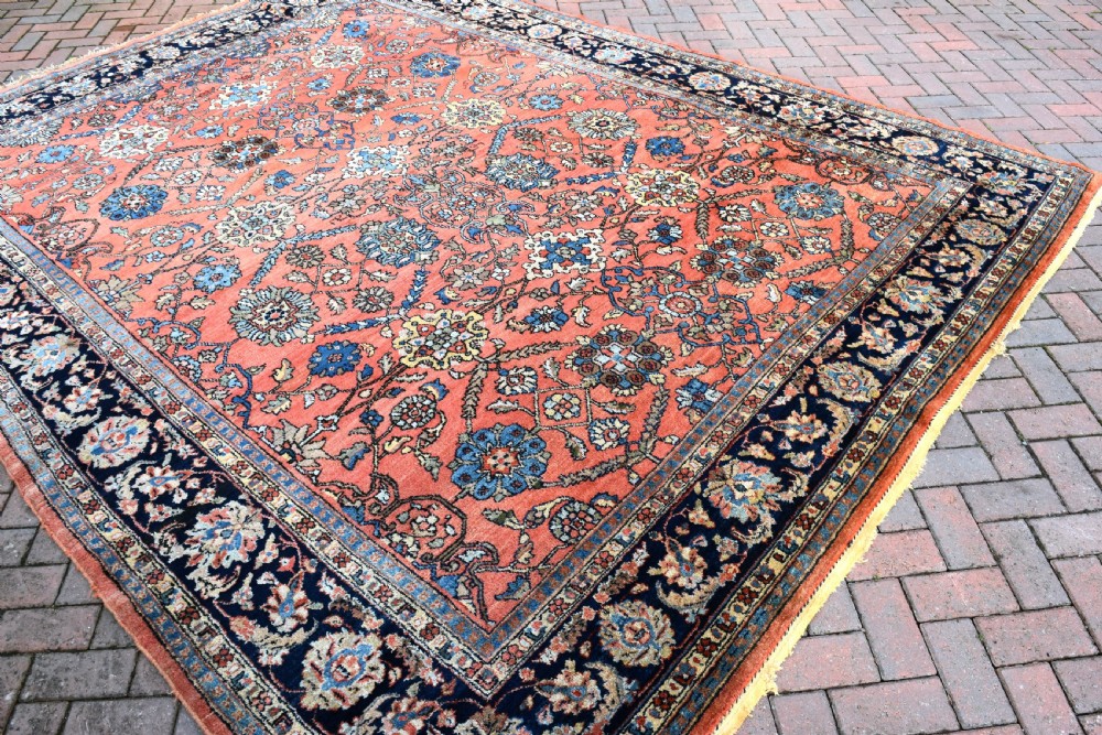 An antique Persian Lilihan wool carpet featuring a terracotta pink field woven with blue floral motifs and framed by a dark navy ornate border. View VII