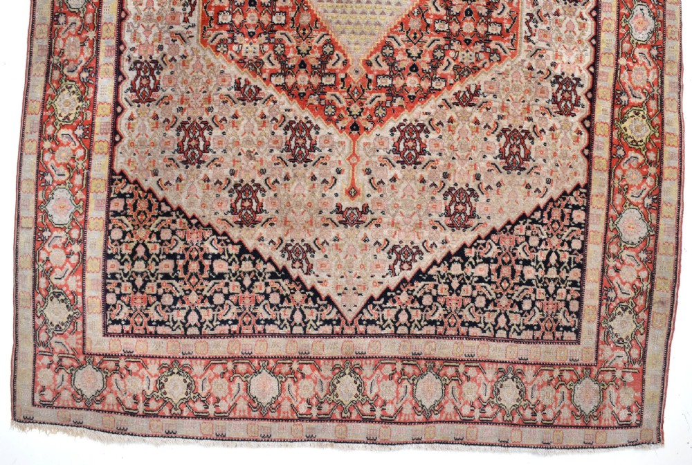 An antique Persian Senneh hand-knotted wool rug featuring a central cream stepped medallion surrounded by intricate red and blue geometric borders. View II