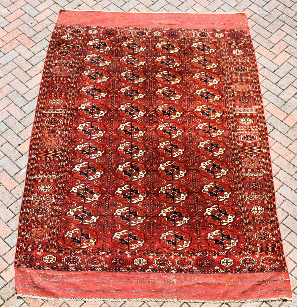 An antique hand-knotted Tekke Turkmen wool main carpet featuring repeating geometric gul motifs on a deep terracotta red field with decorative borders and flatweave ends.