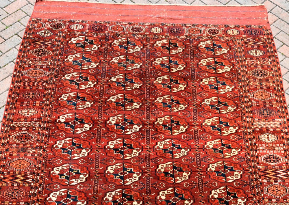 An antique hand-knotted Tekke Turkmen wool main carpet featuring repeating geometric gul motifs on a deep terracotta red field with decorative borders and flatweave ends. View V