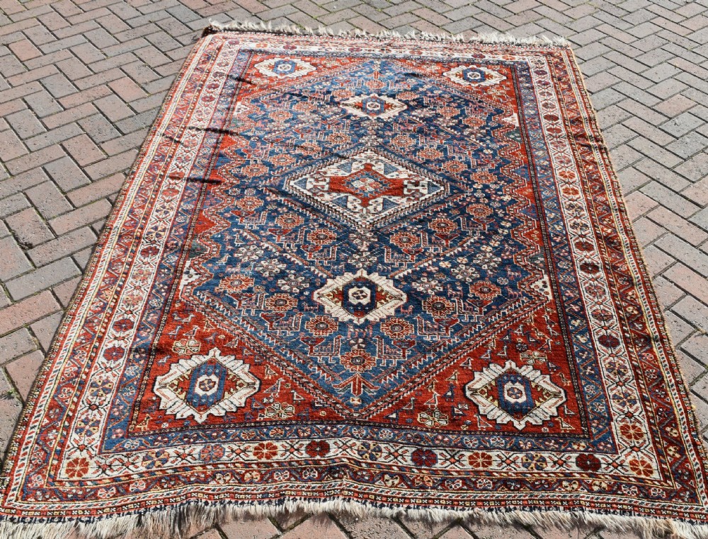 An antique Qashqai tribal wool rug with geometric diamond medallions in navy blue, terracotta red, and cream with fringed edges.