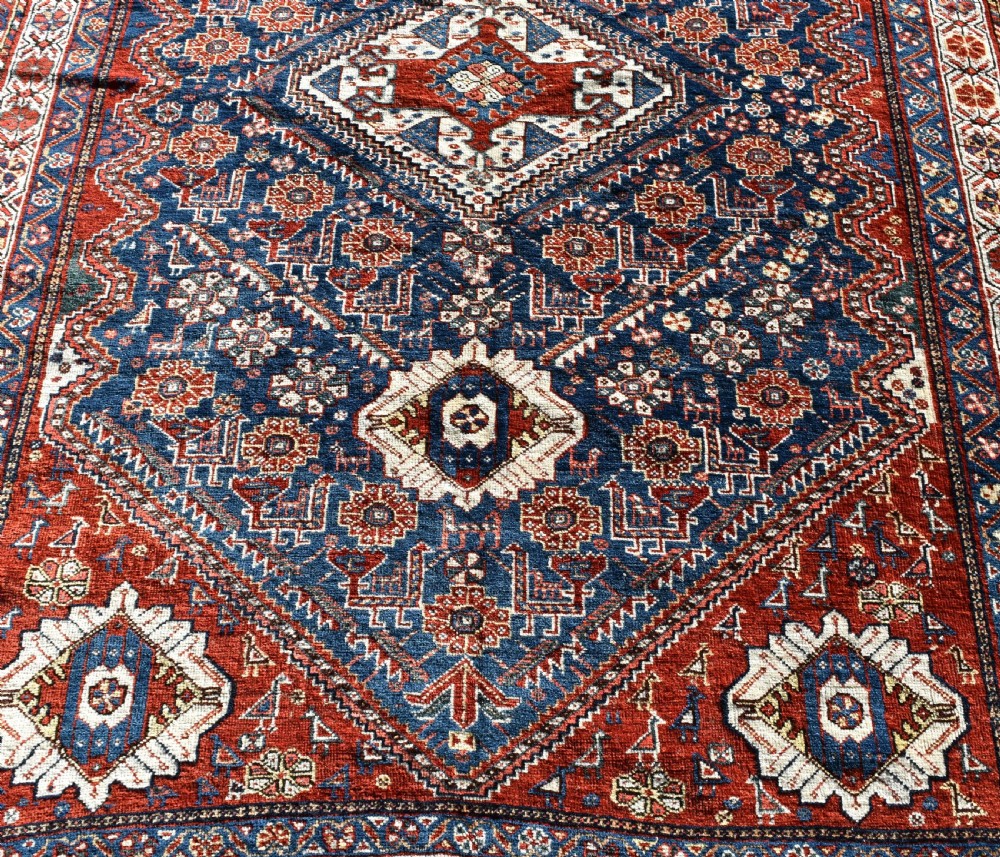 An antique Qashqai tribal wool rug with geometric diamond medallions in navy blue, terracotta red, and cream with fringed edges. View IV