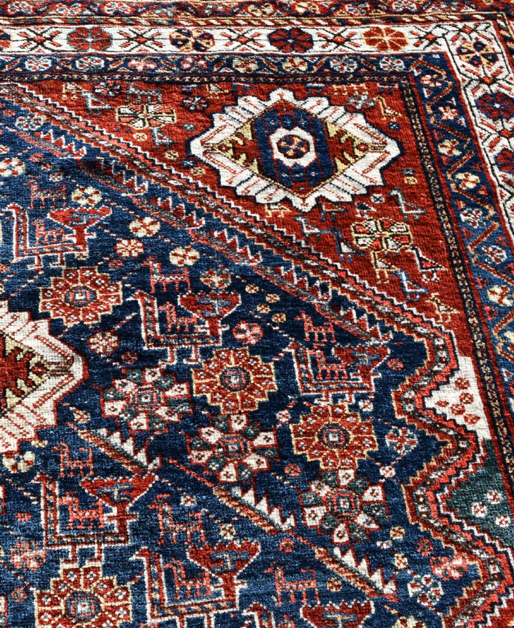 An antique Qashqai tribal wool rug with geometric diamond medallions in navy blue, terracotta red, and cream with fringed edges. View V