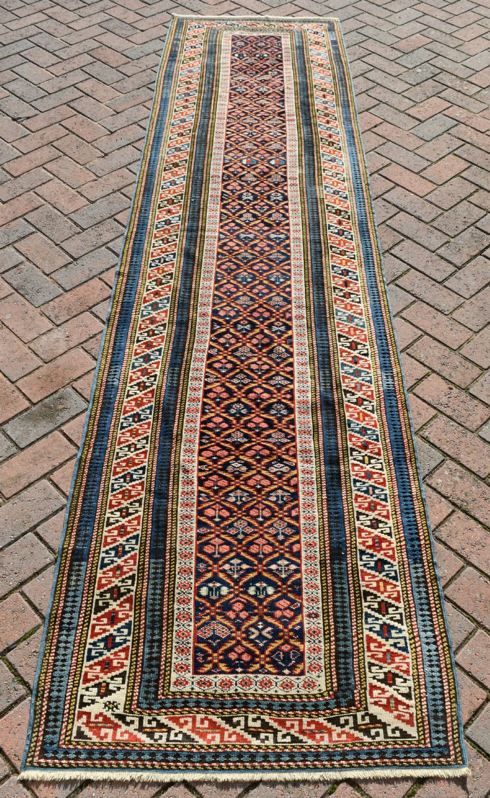A long, narrow hand-knotted antique Caucasian Kuba runner rug featuring a navy blue geometric diamond lattice pattern framed by intricate multi-coloured borders.